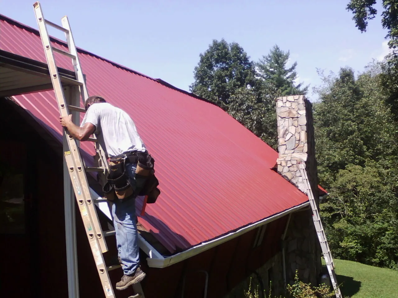 Roofer on a ladder beside a red metal roof and stone chimney — Soffit & Fascia Repair by Roofing Brothers in Steinhatchee
